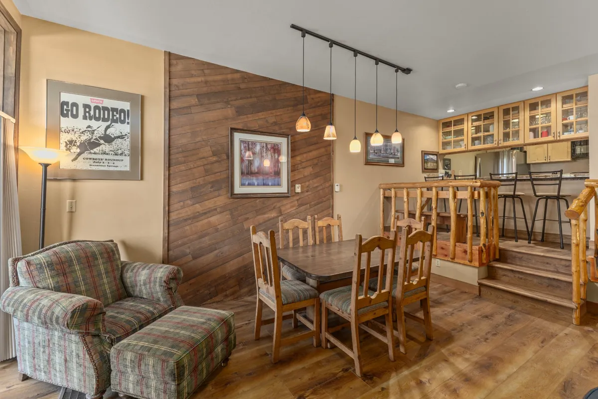 A warm dining area with a wooden table, plaid armchair, and ambient pendant lighting, set beside a wooden staircase and framed artwork. The space is ideal for group gatherings, game nights, or shared meals.