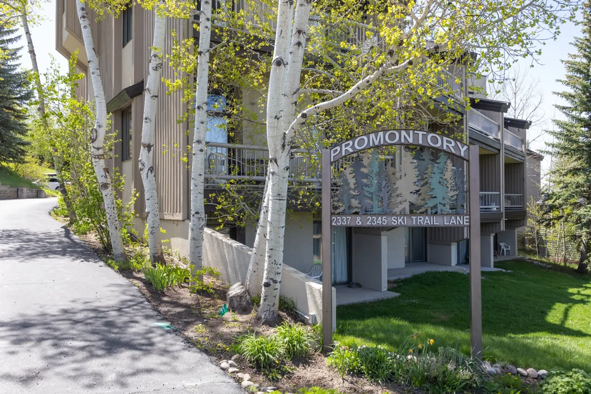 This exterior shot captures the building's welcoming facade, with signage, surrounding trees, and a pathway inviting guests to step outside and explore. The scene is set in a winter landscape, conveying proximity to ski trails and the mountain environment.
