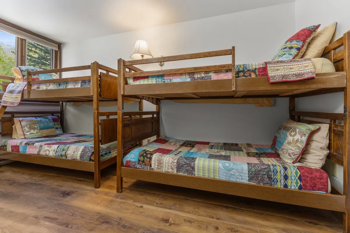 A bright bunk bedroom featuring two sets of bunk beds adorned with colorful quilts. Natural light streams in through the window, enhancing the playful and inviting atmosphere. The space appears organized and ideal for children or singles.