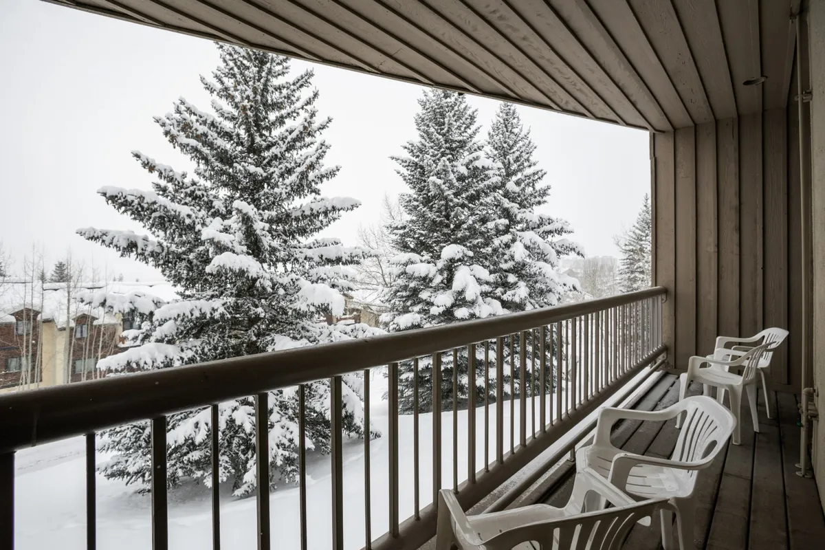 A serene winter balcony scene featuring snow-covered trees, a balcony railing, and empty chairs. The setting suggests a peaceful retreat, ideal for enjoying a warm drink after a day on the slopes or watching the snow fall in quiet reflection.