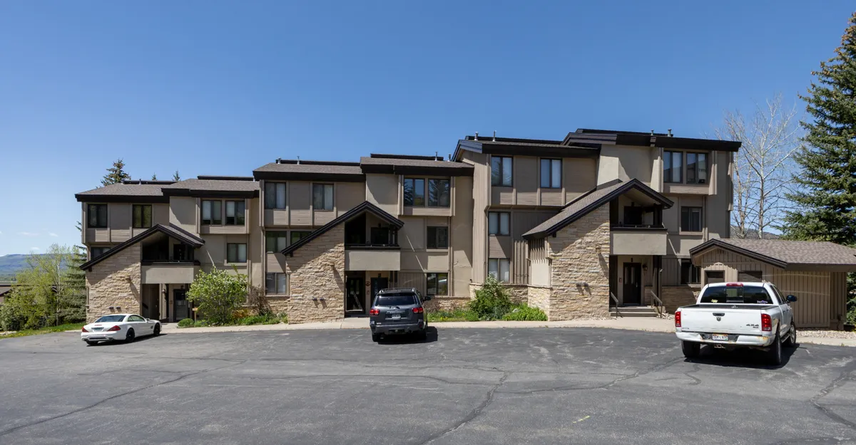 A wide exterior view of the multi-story building with a stone facade, surrounded by landscaped greenery and a parking area. The inviting and tranquil mood of the scene suggests a welcoming base for all your summer adventures, while also hinting at the ease of access to local events and the neighborhood.
