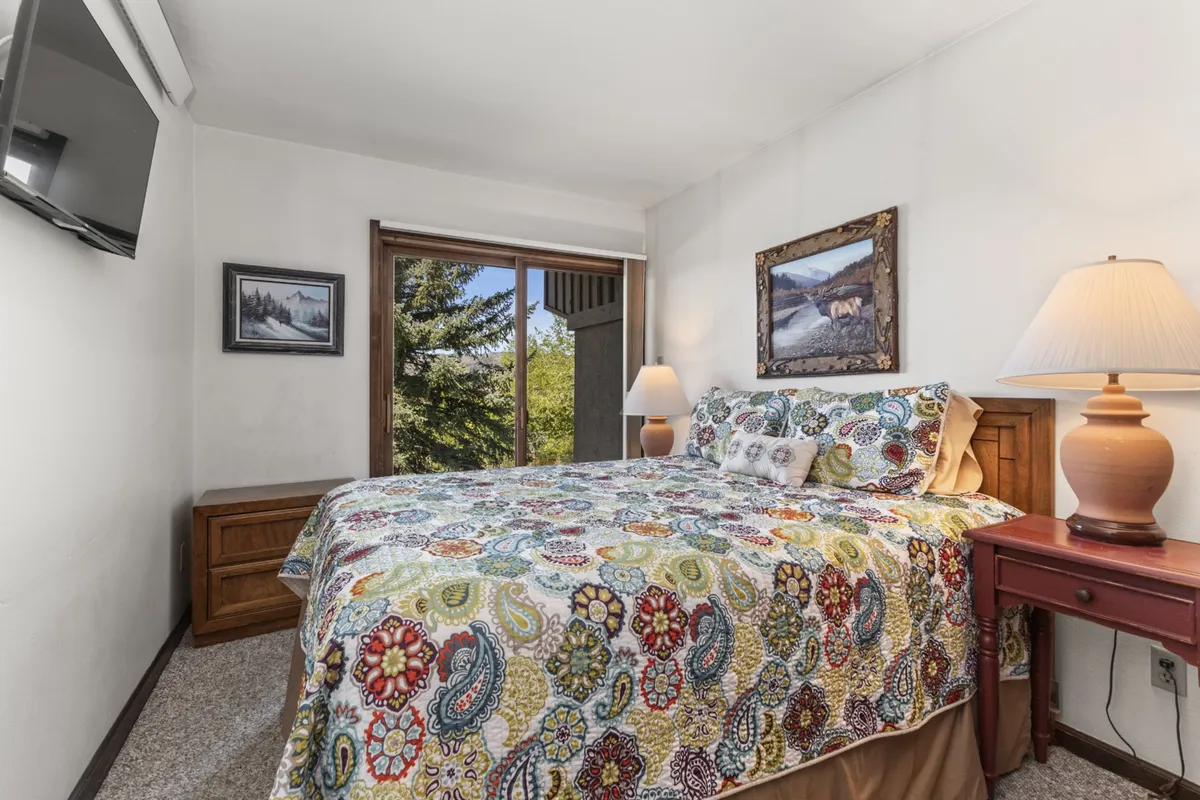A cozy queen bedroom with a colorful bedspread, nightstand with a lamp, framed artwork, and a large window overlooking green trees. The setting is inviting, with soft natural light creating a restful atmosphere.