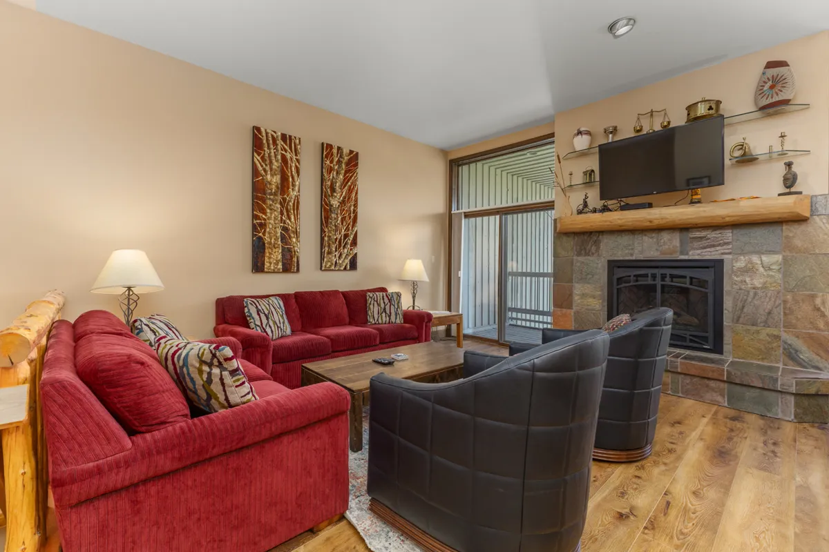 This inviting living area features plush red sofas and black armchairs arranged around a stone fireplace, with wooden shelves lining the walls. The space feels cozy yet airy, suggesting both comfort during winter evenings and the option to open doors for summer breezes.