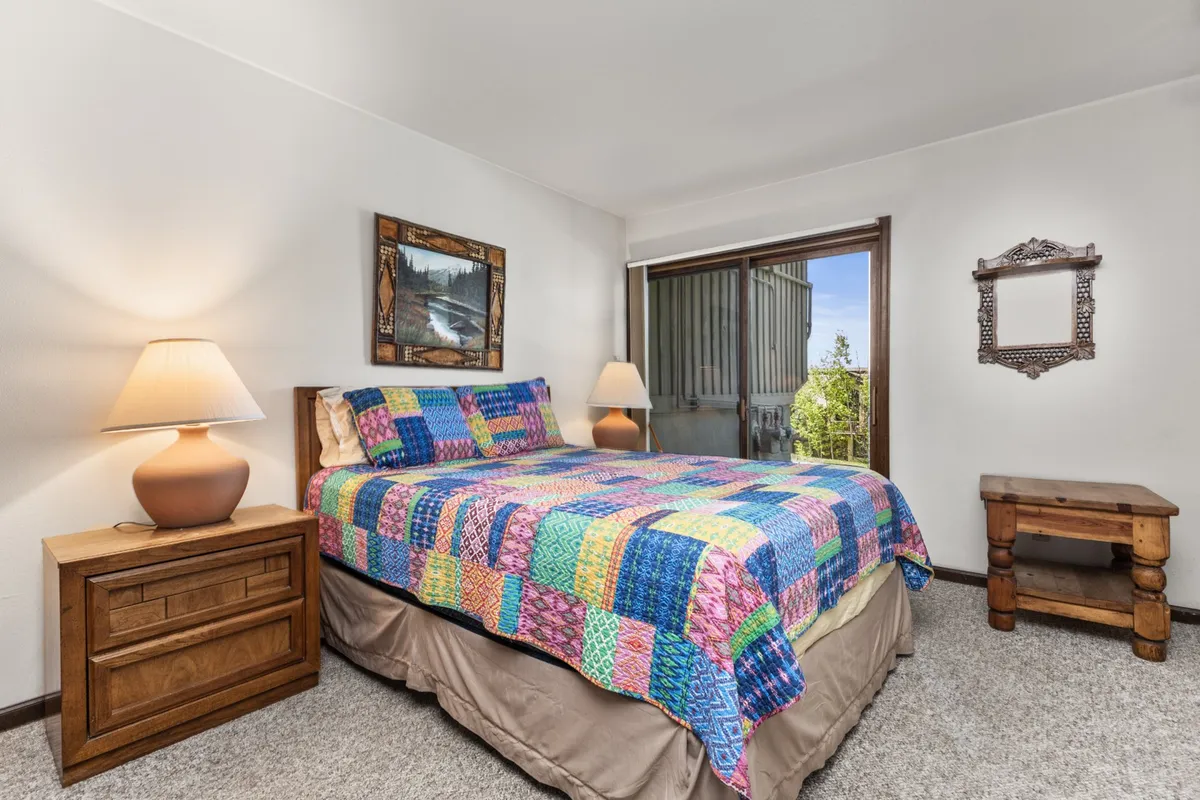 A welcoming bedroom with a queen-sized bed covered in a vivid quilt, a wooden nightstand and lamp, landscape artwork, and a large window bringing in sunlight. The atmosphere is cheerful and perfect for relaxation or reading.