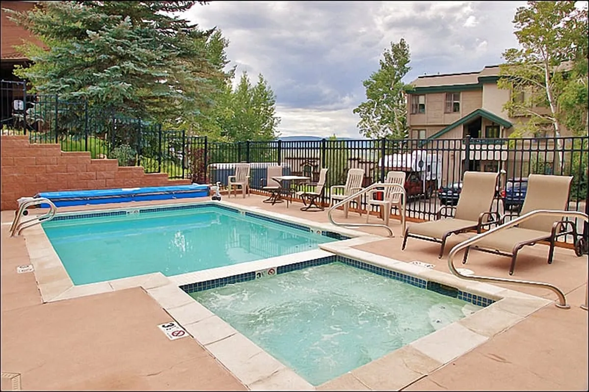The image showcases the condo's outdoor amenities, with a sparkling swimming pool and inviting hot tub surrounded by lounge chairs and lush greenery—perfect for relaxation or socializing after a day on the slopes.